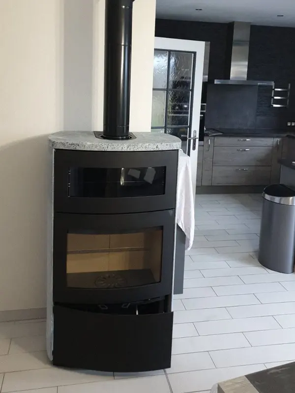 Poêle à bûches noir avec compartiment supérieur vitré et dessus en granit, près d'une cuisine