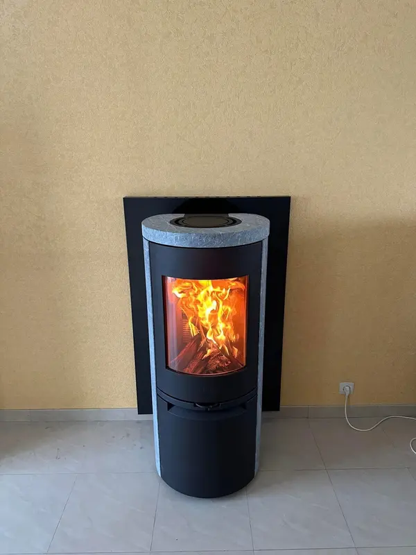 Poêle à bûches rond avec dessus en pierre ollaire, flambée allumée, devant une plaque de protection murale
